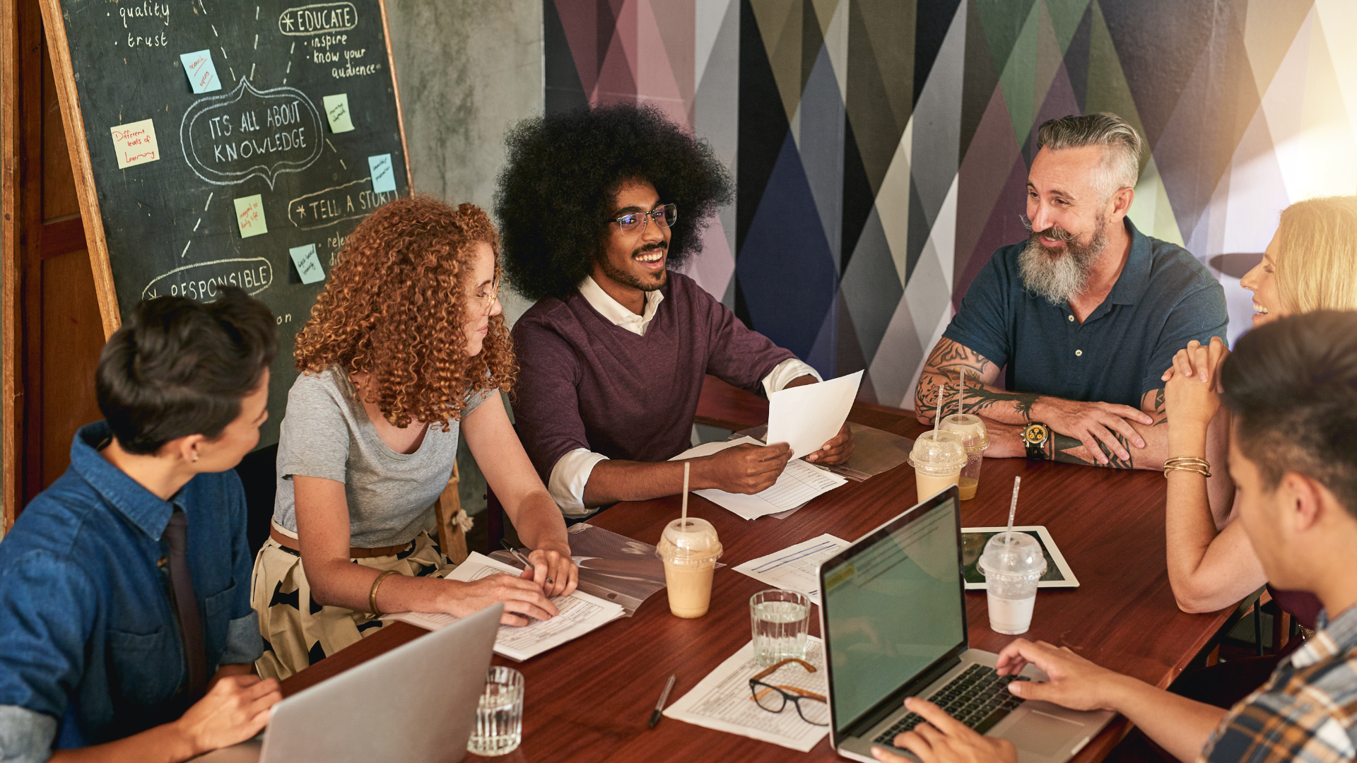 6 people sitting around a table with a chalk board in the background collaborating and having a meeting.