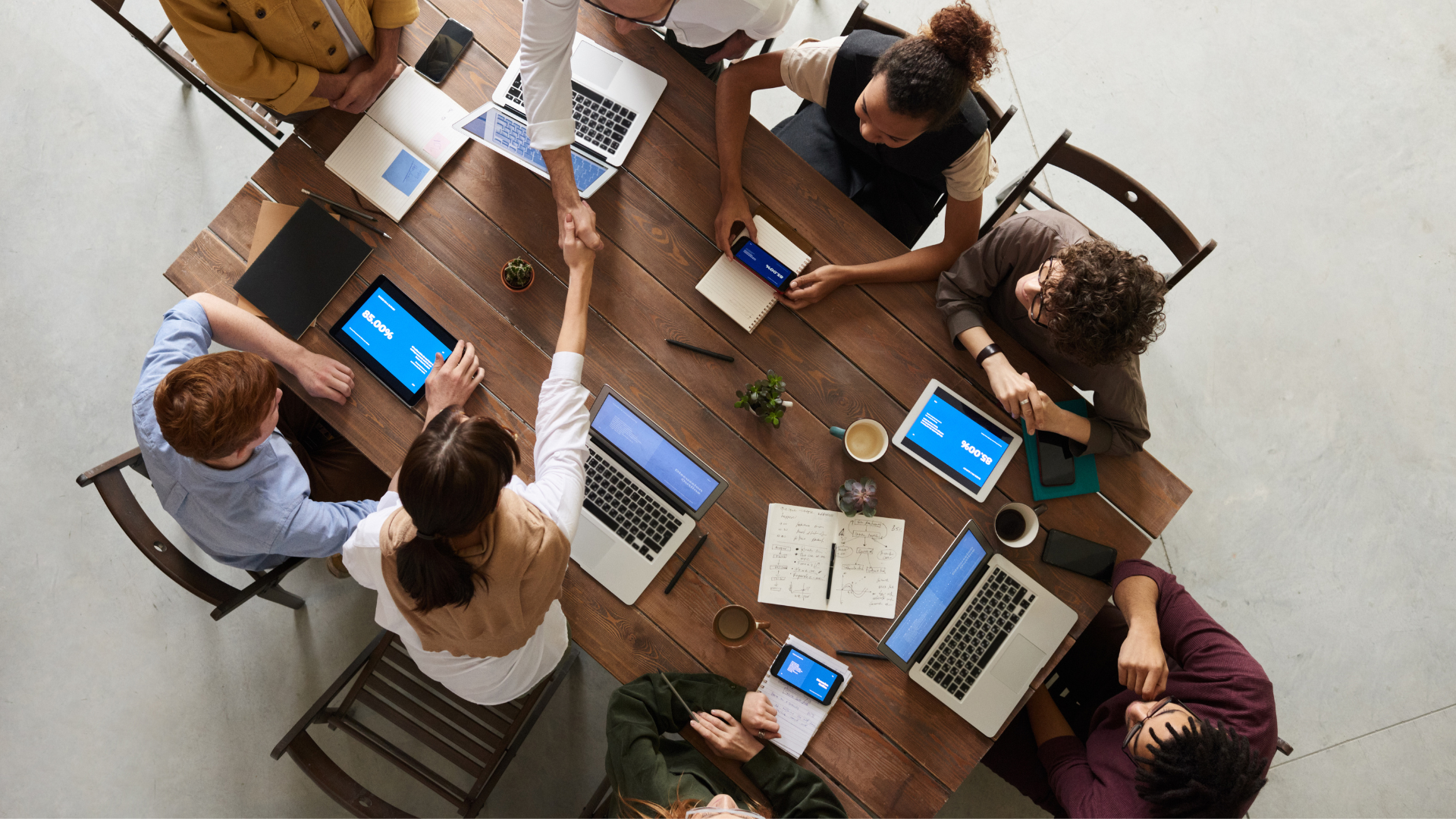 Team of 8 sitting around a table with their laptops shaking hands.