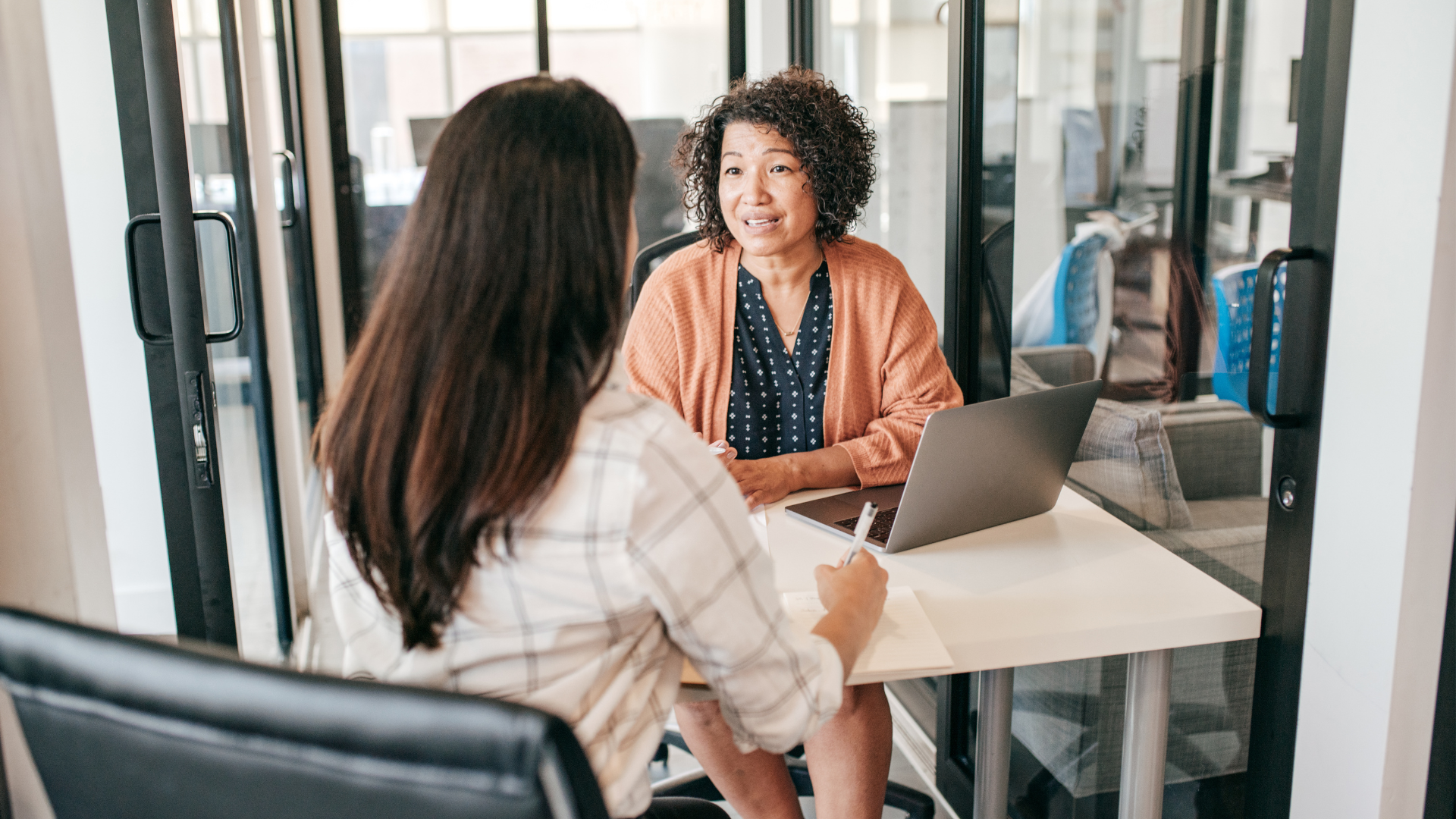 Woman with curly hair sitting behind a desk and laptop interviewing another woman