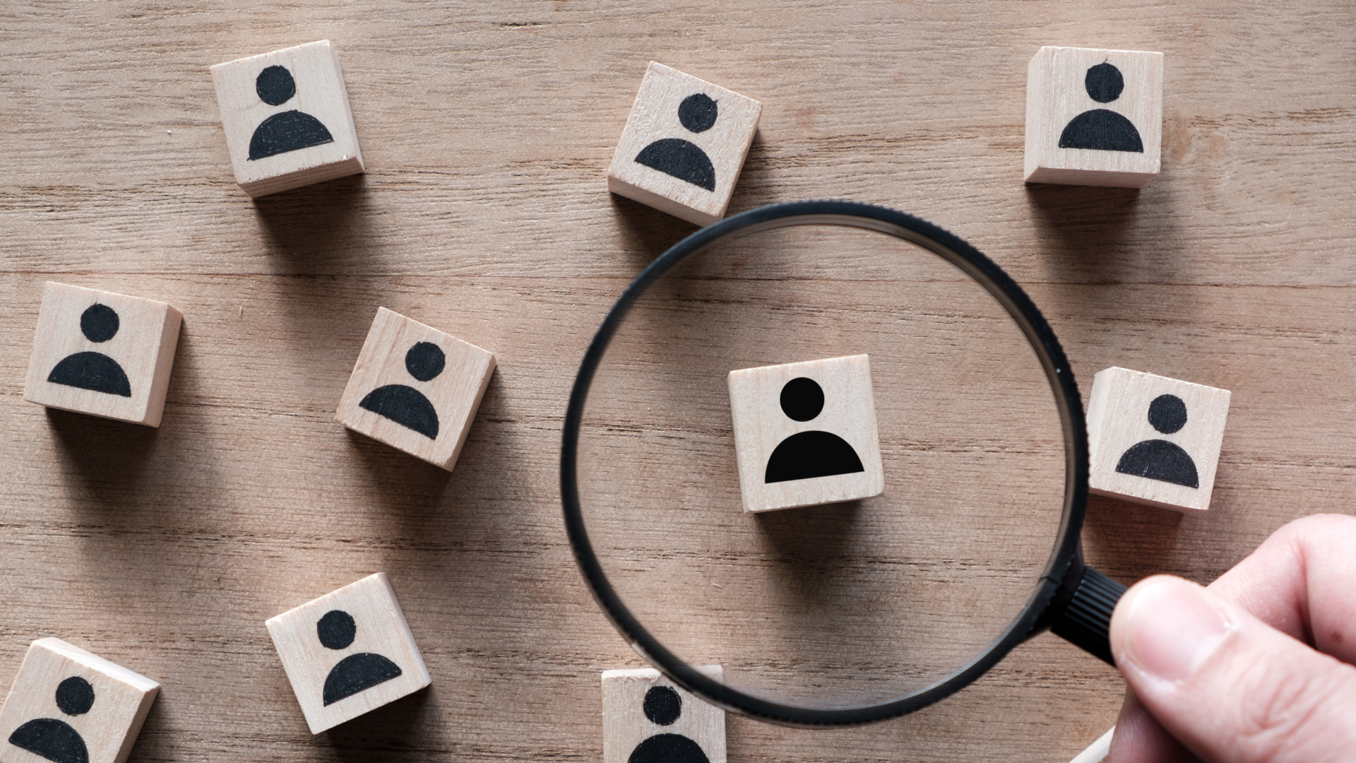 Small wooden squares with people graphic on them under a magnifying glass sitting on a wooden table.